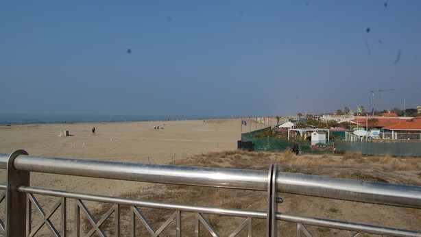 A beach with a metal railing in the foreground, a sandy beach, and buildings in the background. - VERSILIA APARTMENT (Pietrasanta)