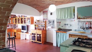 A kitchen with wooden cabinets, a gas stove, and a dining area with a chair and table. - The Photographer's House (Colle di Val d'Elsa)