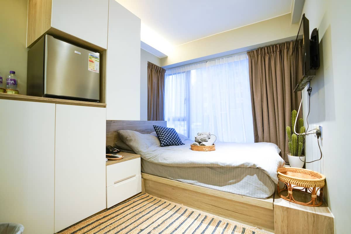 Un dormitorio pequeño con cama, mesita de noche, televisor integrado y ventana con cortinas.
