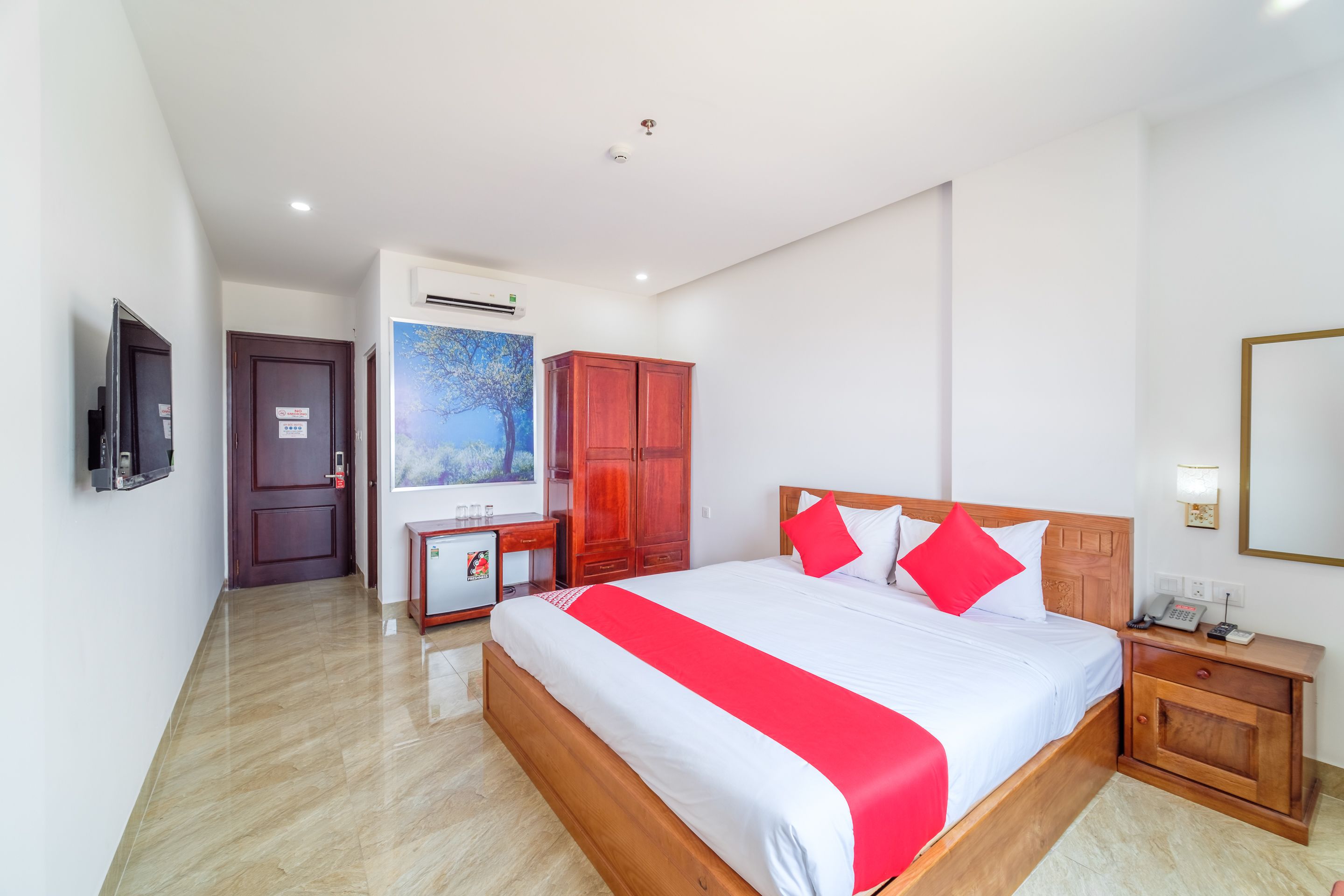 A hotel room with a large bed, wooden wardrobe, a desk with a chair, a TV mounted on the wall, and an air conditioning unit.