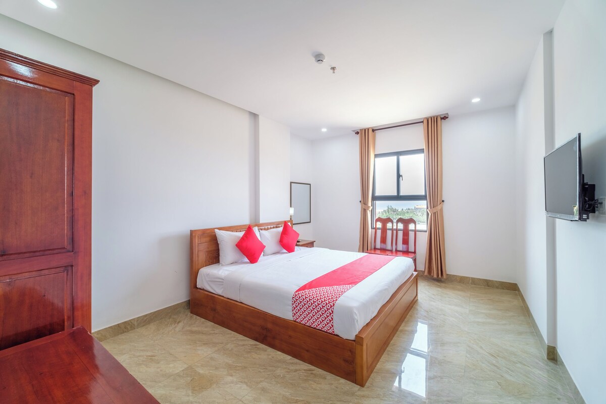 Une chambre d’hôtel avec un lit en bois, des murs blancs, une télévision et un balcon avec des rideaux.