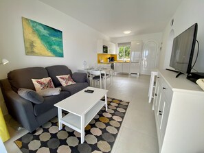 Un soggiorno moderno con un divano grigio, un tavolino da caffè bianco e un tappeto colorato. Sullo sfondo, si intravede una cucina con mobili bianchi e un'area pranzo.