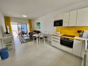 Cucina moderna con mobili bianchi, un forno a microonde e un tostapane. C'è un'area pranzo con tavolo e sedie, e vista sulla spiaggia attraverso la porta scorrevole in vetro.