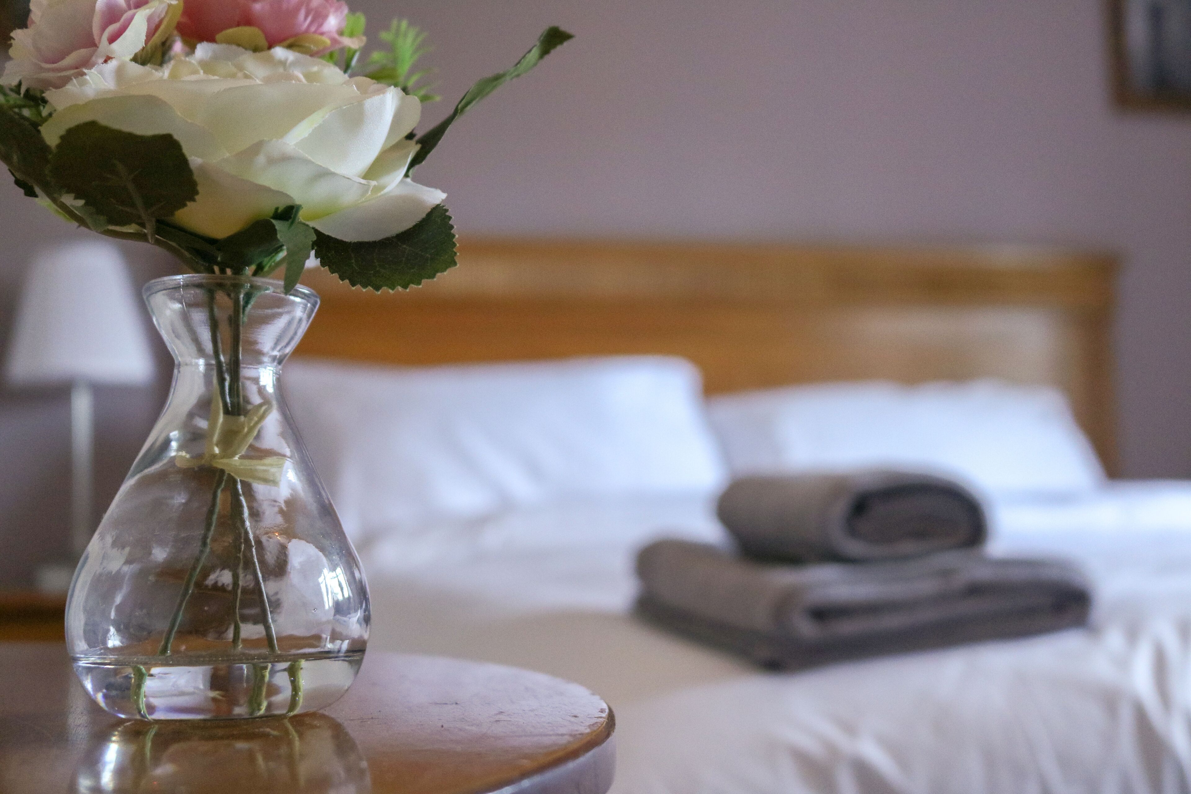 A clear glass vase with flowers on a bedside table in a hotel room.