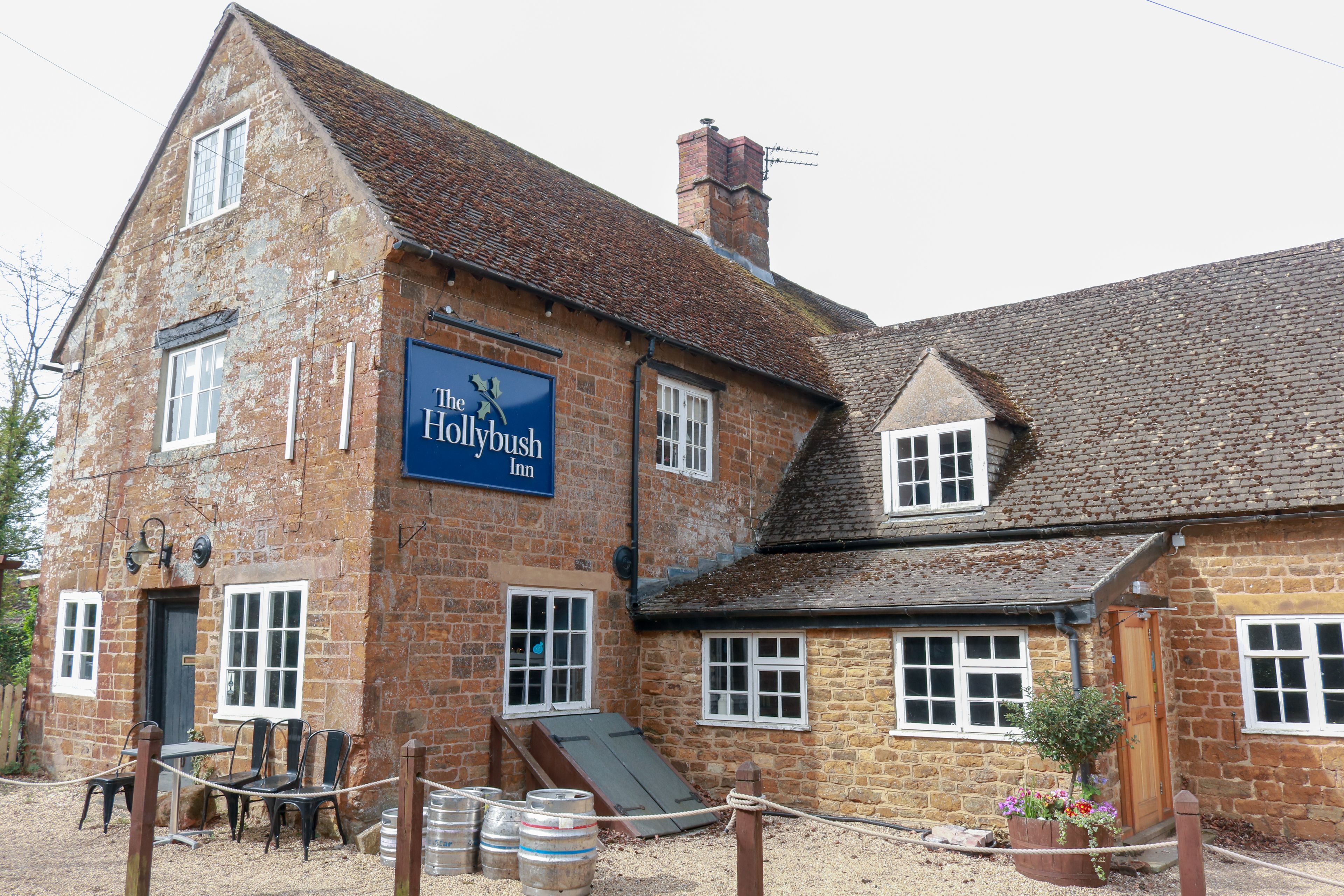 A traditional brick building with a sign reading "The Hollybush Inn." It features a small outdoor seating area with tables and chairs, a wooden door, and a window with a white frame.