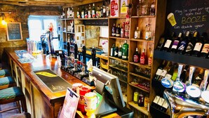 A bar with shelves of liquor bottles, a cash register, and a chalkboard with writing. - The Hollybush Inn (Southam)