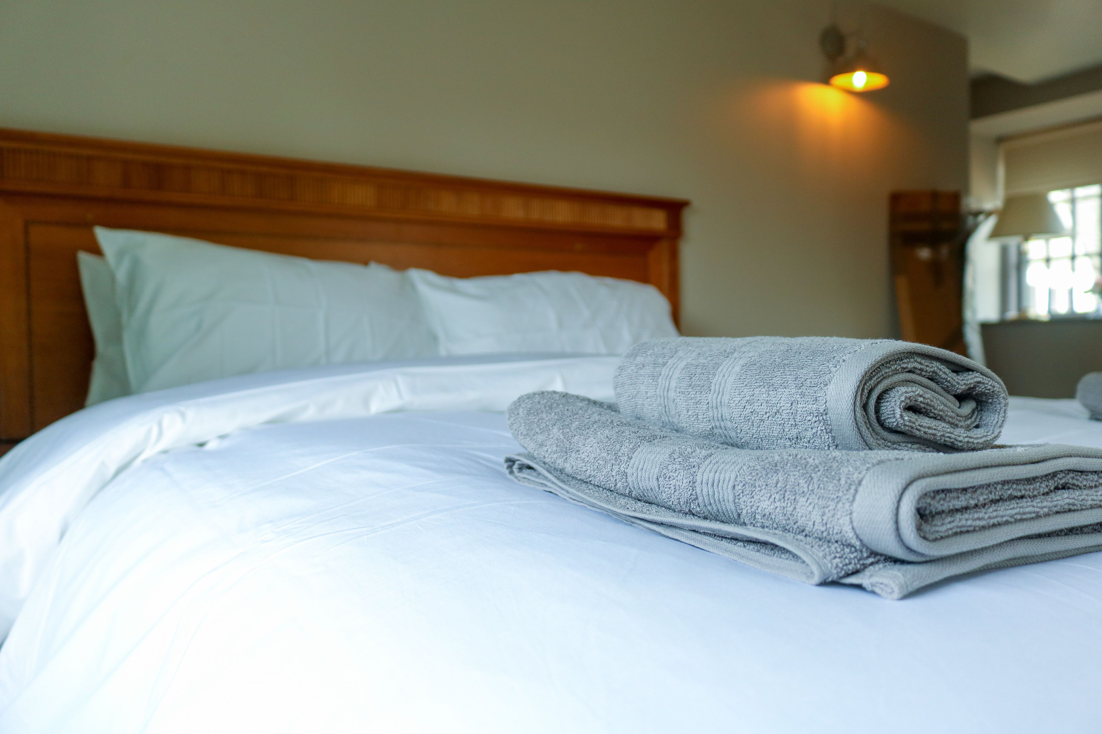 A neatly made bed with white linens and a rolled gray towel, a wooden headboard, and a wall-mounted light fixture.