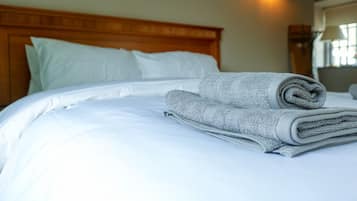 A neatly made bed with white linens and a rolled gray towel, a wooden headboard, and a wall-mounted light fixture.