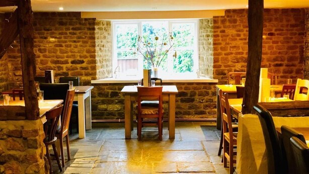 A stone-floored dining area with tables and chairs, a window with a view of greenery, and a rustic interior. - The Hollybush Inn (Southam)