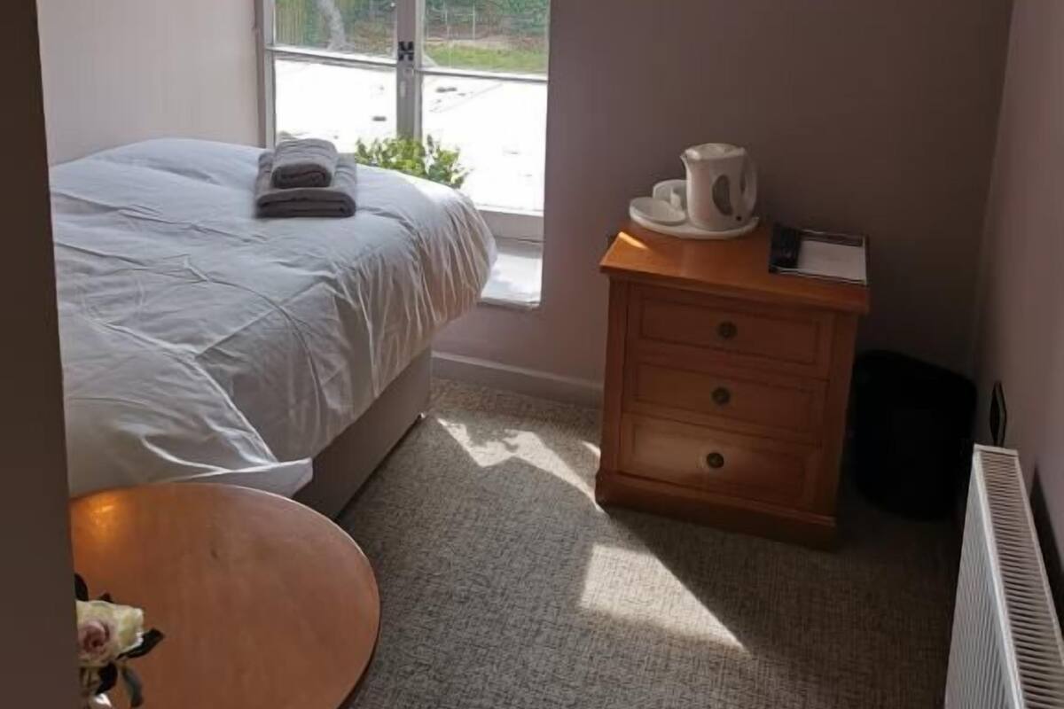 A bedroom with a bed, a wooden nightstand with a kettle, a small table with a vase, and a window with a view of greenery.