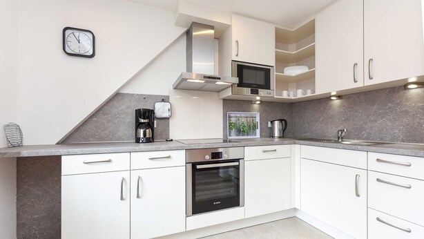 A modern kitchen with white cabinets, a Bosch oven, a microwave, and a clock on the wall. - Apartment top right - House Albatros (Neuharlingersiel)