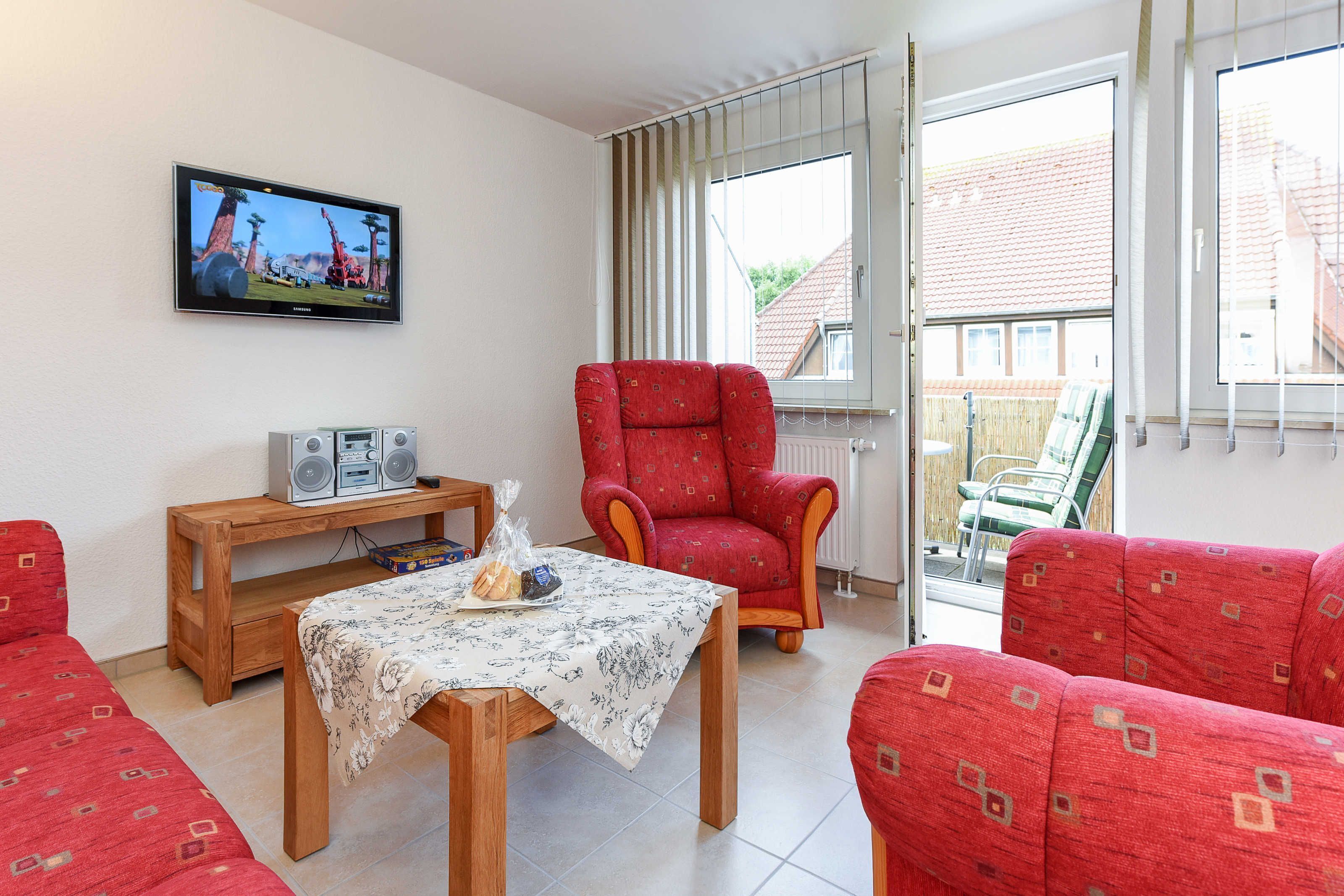 Een woonkamer met een televisie, een rode fauteuil, een houten tafel met een bloemrijk tafelkleed en rode gestreepte banken.