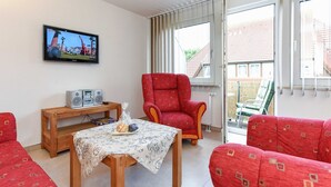 A living room with a television, a red armchair, a wooden table with a floral tablecloth, and red patterned sofas. - Apartment top right - House Albatros (Neuharlingersiel)