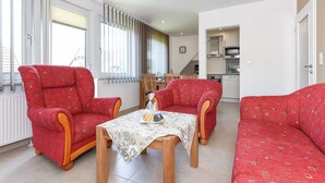 A living room with red patterned sofas, a wooden coffee table with a floral tablecloth, and a kitchen in the background. - Apartment top right - House Albatros (Neuharlingersiel)