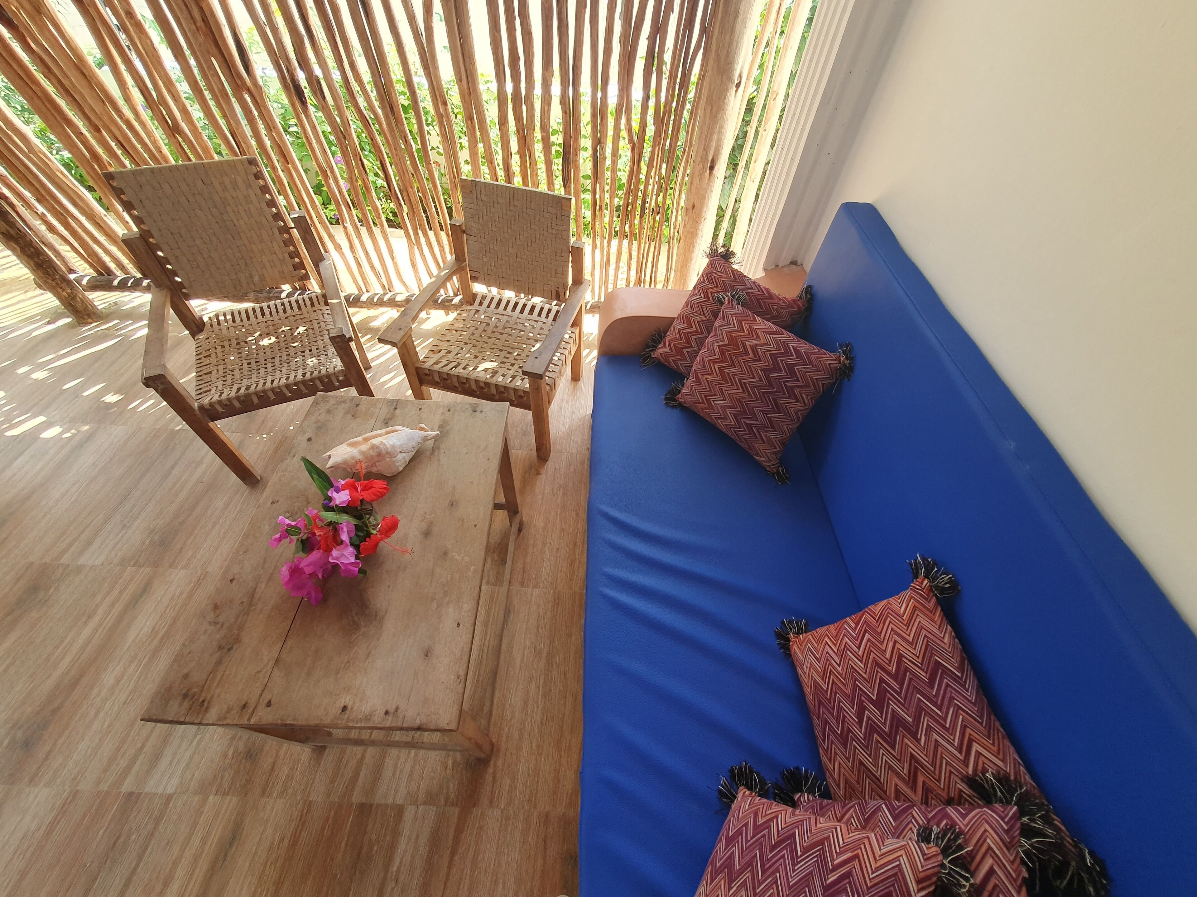 A room with a wooden coffee table, two wicker chairs, a blue sofa with patterned cushions, and a view of greenery through bamboo blinds.