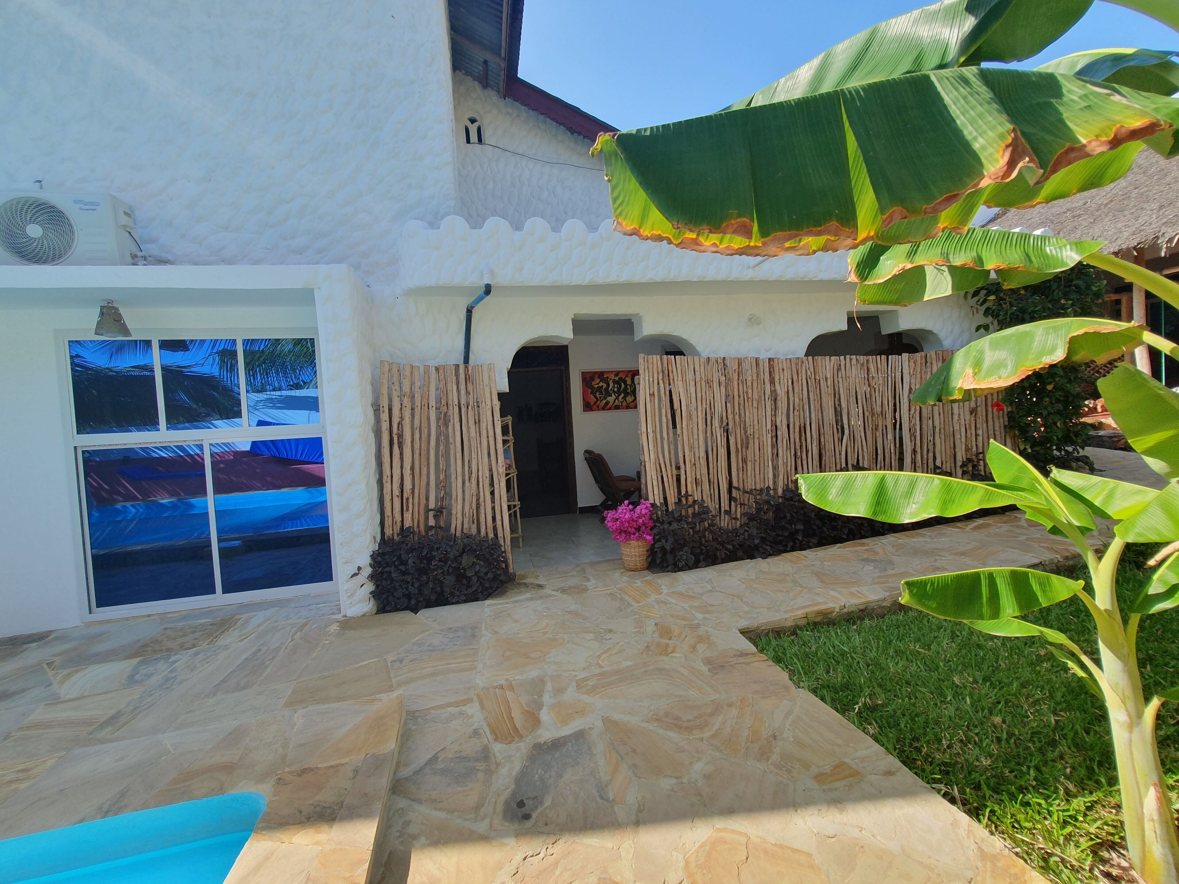 A stone-paved courtyard with a white building, a wooden fence, and a banana plant.