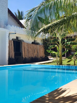 A swimming pool with a wooden deck, a white building with a thatched roof, and a palm tree. - Baridi Villa (Kiwengwa)