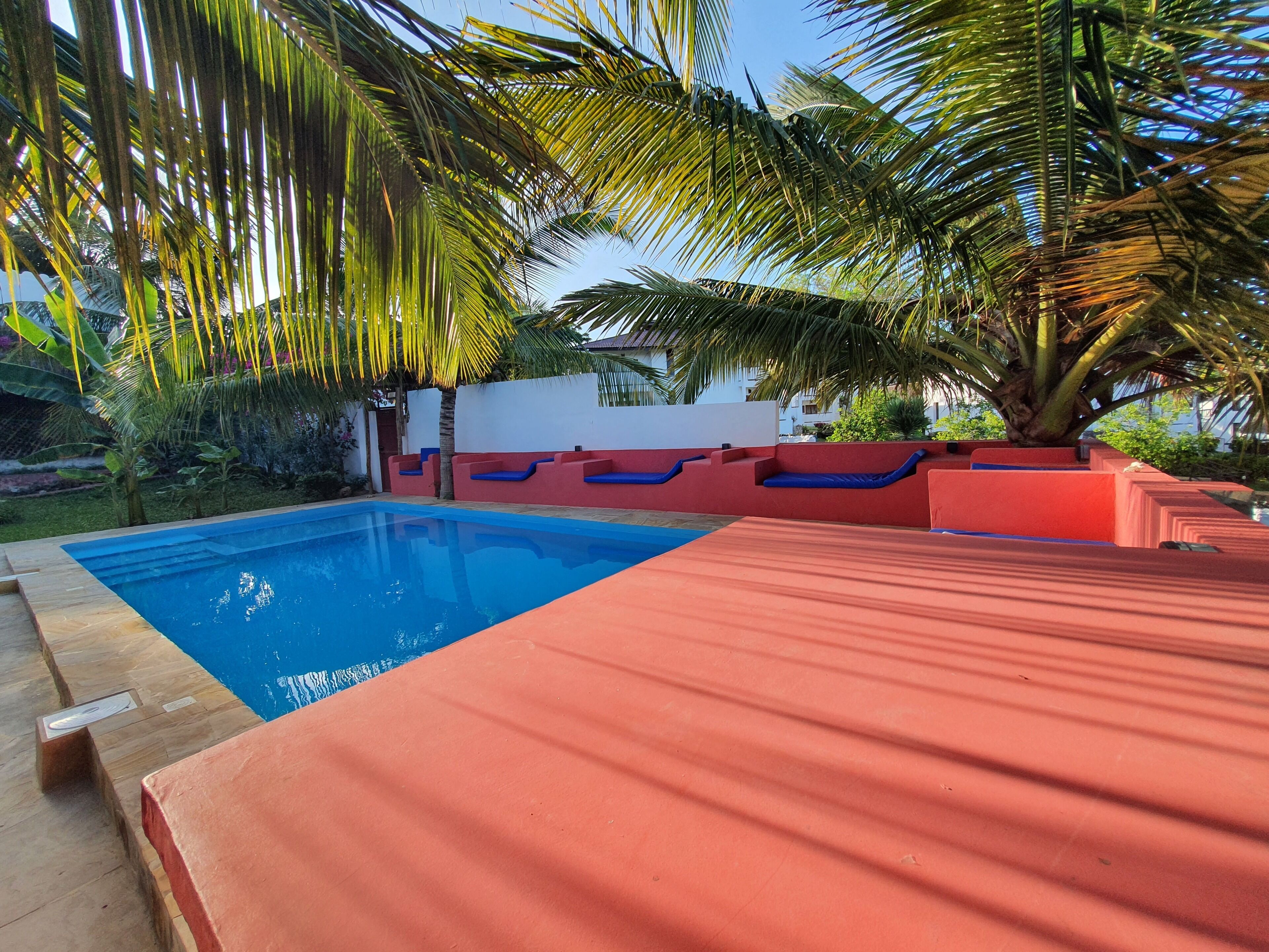 A swimming pool with a red and blue deck, surrounded by palm trees and a white building in the background.