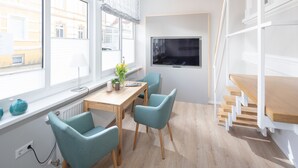 Ein modernes Zimmer mit Holztisch und -stühlen, einem großen Fenster mit Blick auf Gebäude, einem an der Wand befestigten Fernseher und einer Holztreppe.