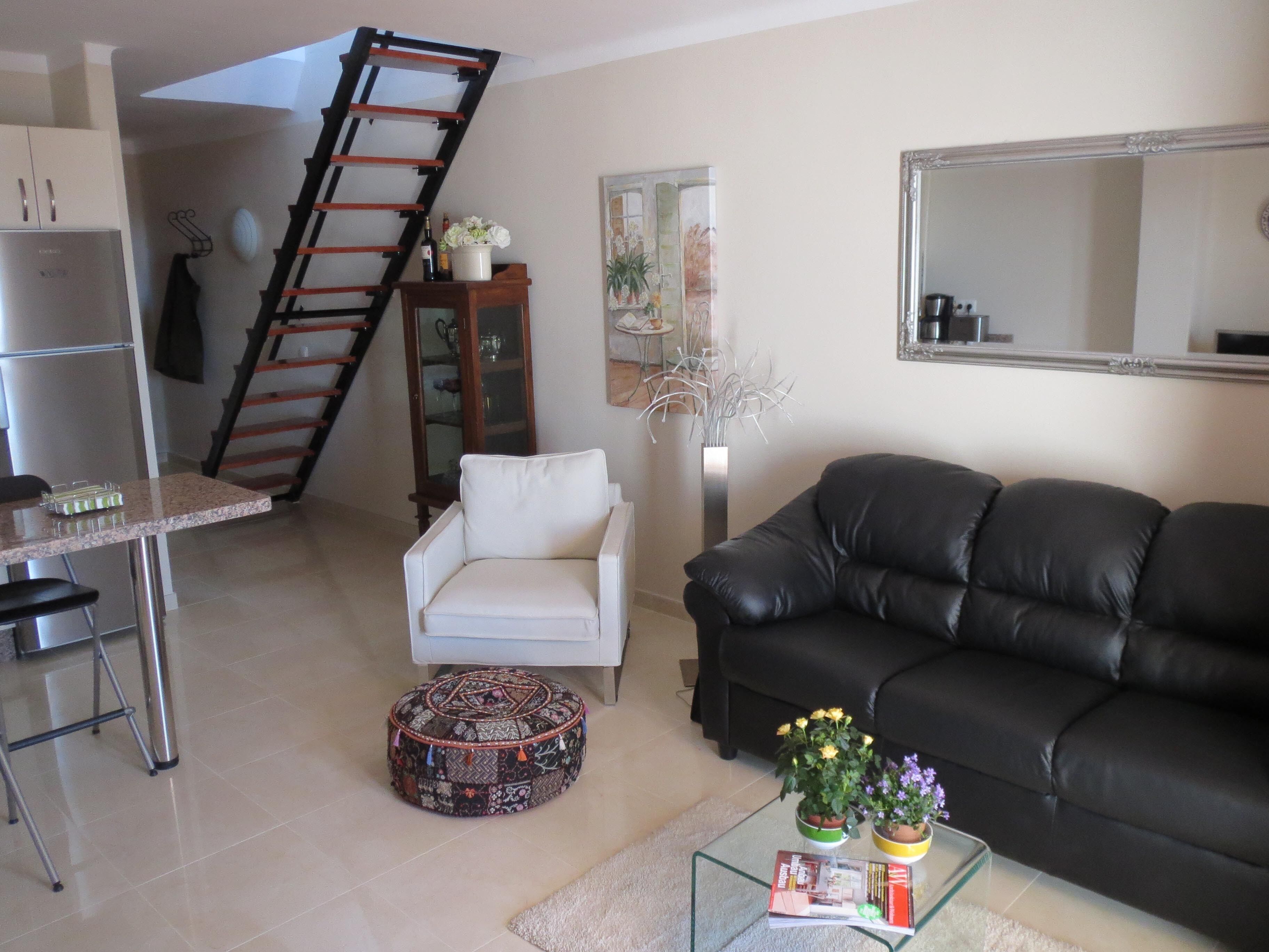 Ein Wohnzimmer mit einer Couch, einem Sessel, einem Couchtisch mit Büchern, einer Leiter und einer Küchenzeile inklusive Kühlschrank.