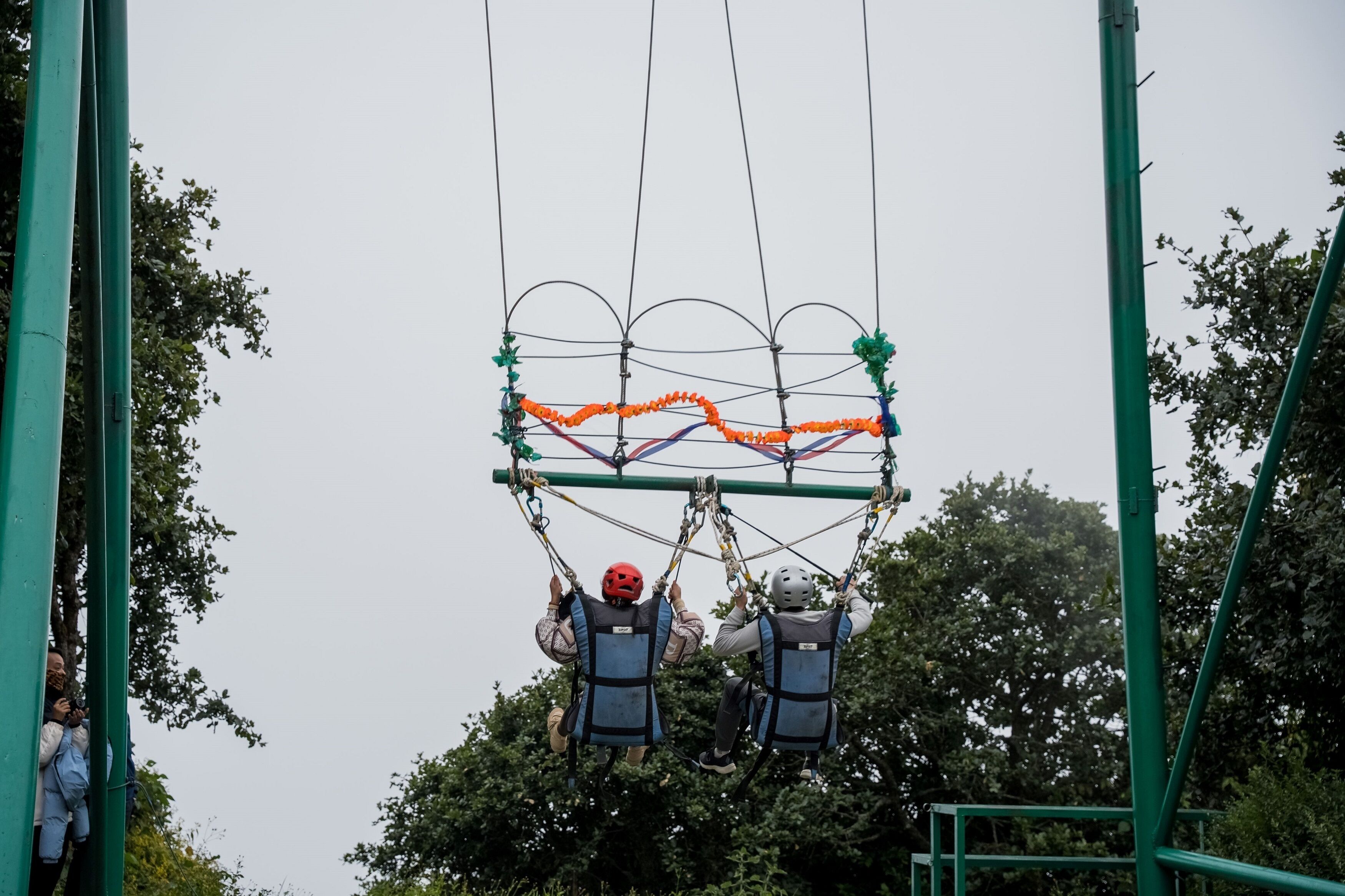 A zip line with two people in safety harnesses and helmets, suspended between two green metal structures.