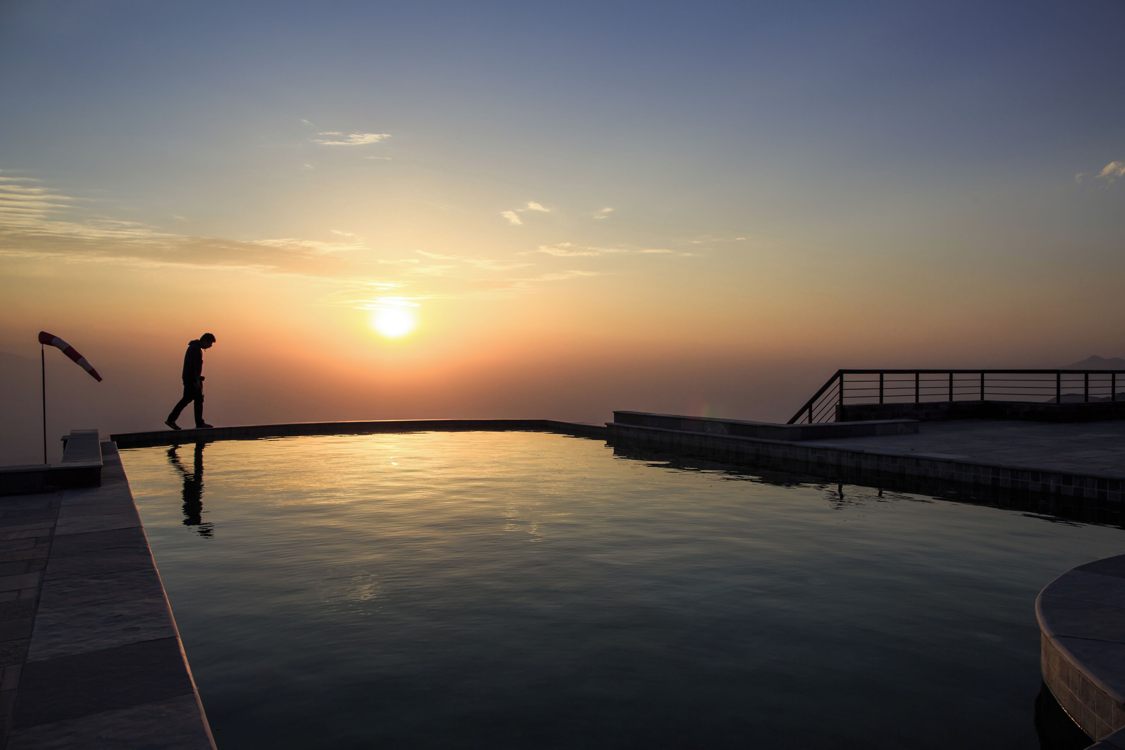 Área de piscina tranquila ao pôr do sol, com uma pessoa caminhando, refletindo a luz do sol na água.