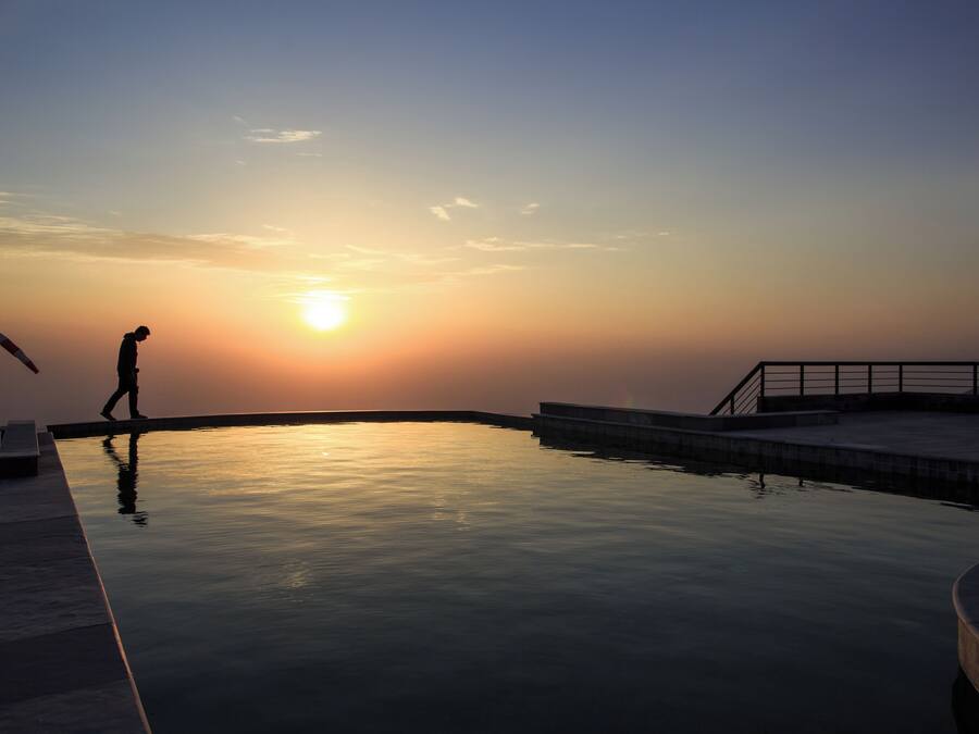 Área de piscina tranquila ao pôr do sol, com uma pessoa caminhando, refletindo a luz do sol na água.