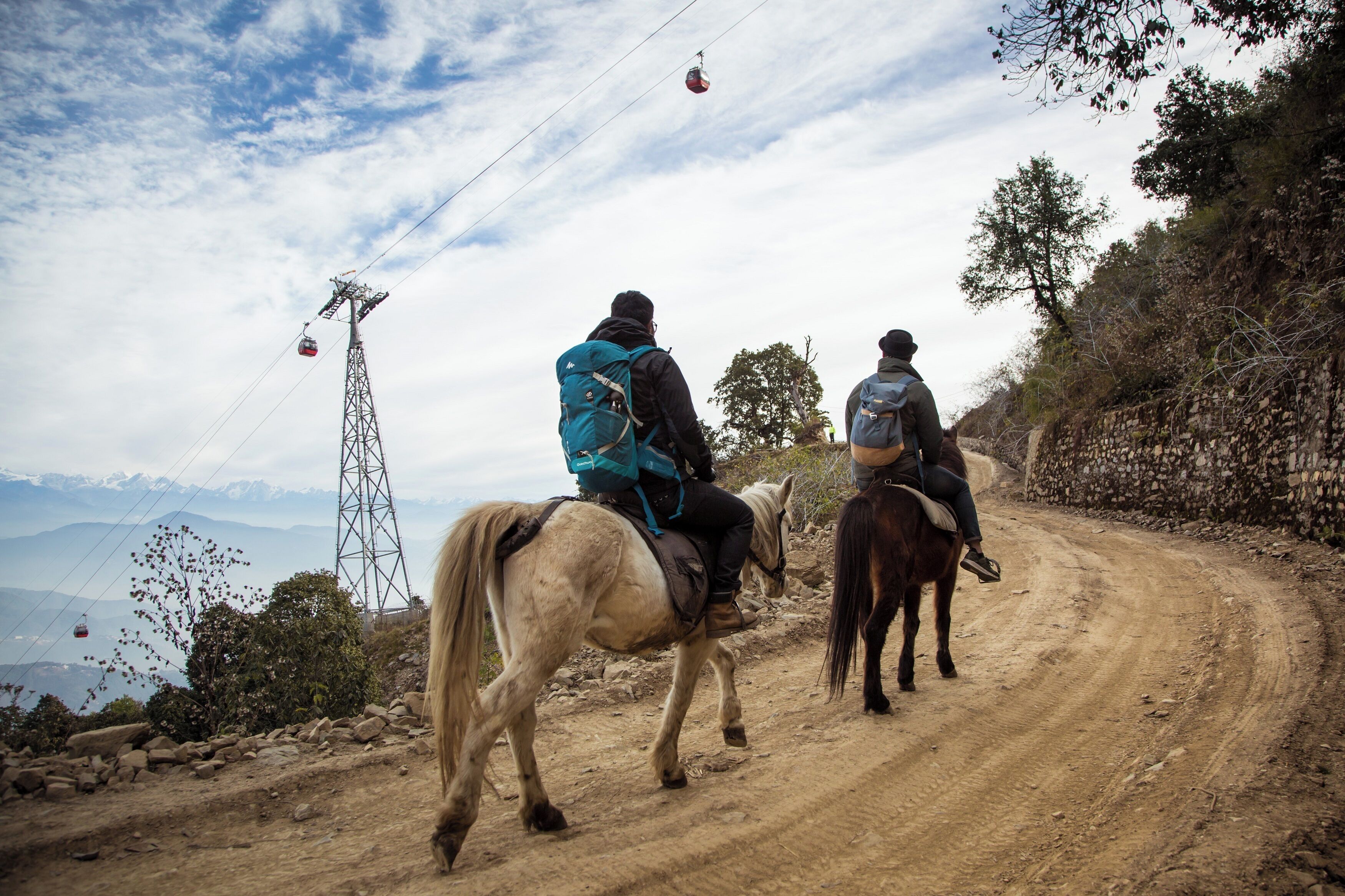 Zwei Personen reiten auf Pferden einen Feldweg entlang, im Hintergrund Berge und ein Strommasten.