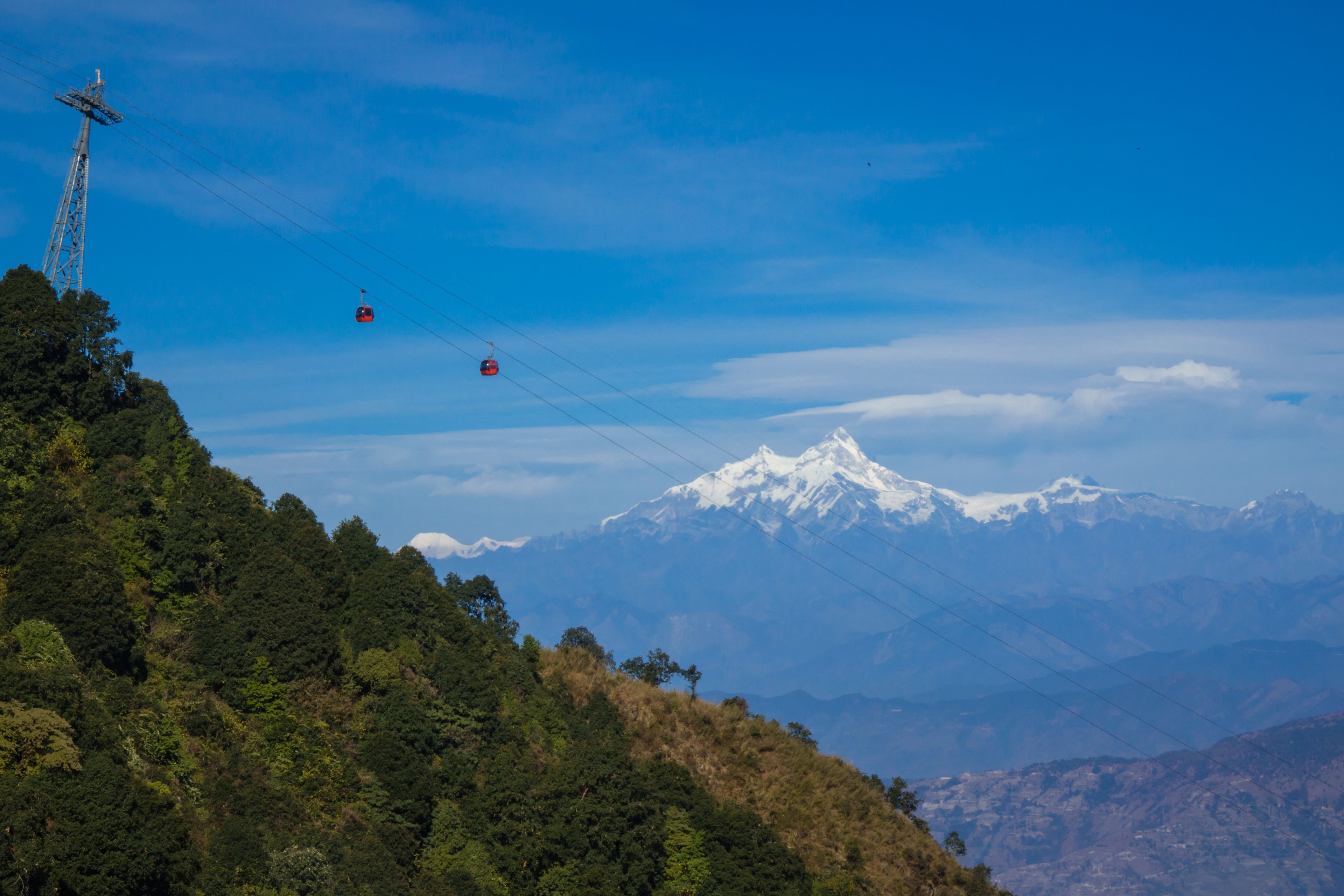 A mountainous landscape with a cable car system, a transmission tower, and a snow-capped peak in the distance.