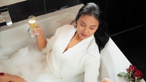 Una persona en una bañera con pétalos de rosa y una copa de champán, con una ventana arriba.