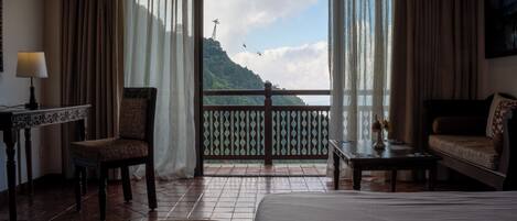 Une chambre d’hôtel avec un lit, une chaise, une petite table et une vue sur un paysage montagneux, avec un téléphérique en vue.