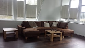 A modern living room with a brown leather sectional sofa, a small wooden coffee table, and a wooden side table. Large windows with blinds allow natural light into the room.