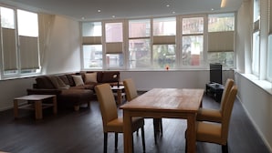 A modern living room with a wooden dining table, cushioned seating, and large windows offering views of the city.