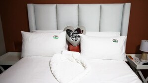 Un lit bien fait, avec du linge de lit blanc et des oreillers, un coussin en forme de cœur portant un message, et une serviette posée sur le lit.
