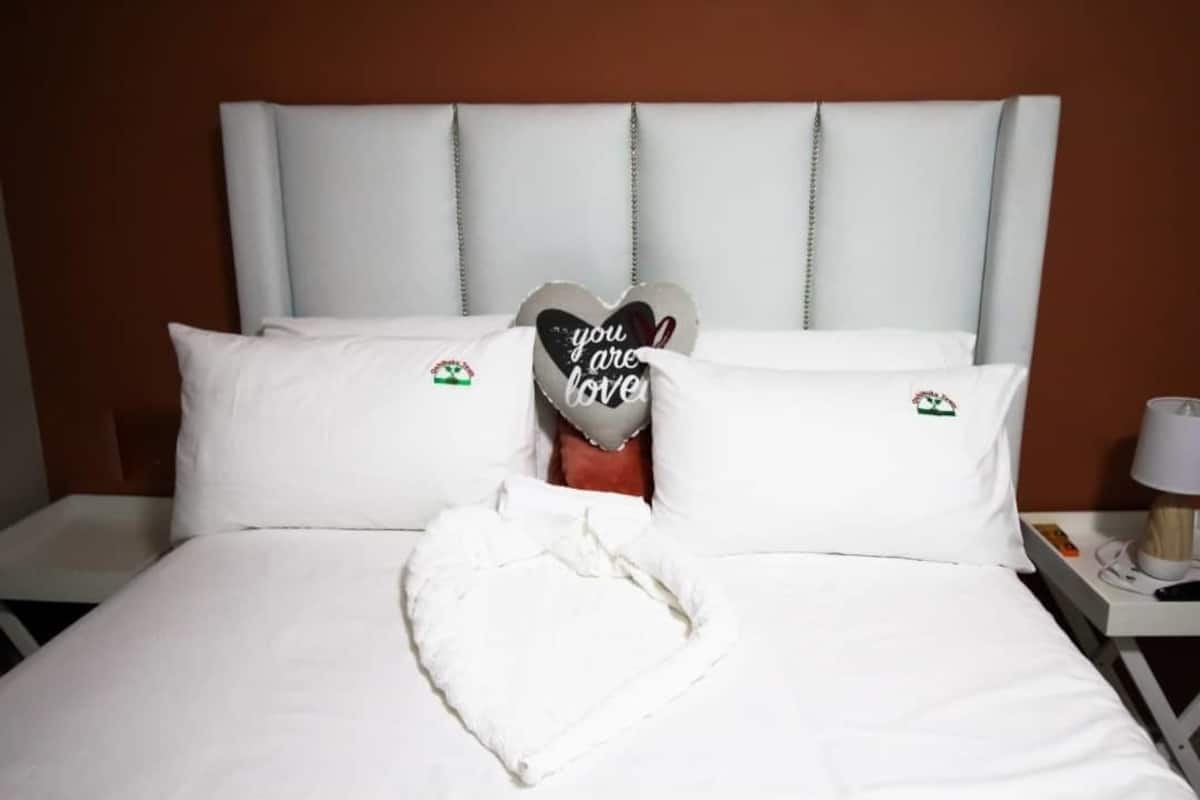 Una cama bien hecha con sábanas y almohadas blancas, incluyendo una almohada con forma de corazón y un mensaje, además de una toalla sobre la cama.