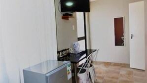 Une pièce de taille modeste équipée d’un petit réfrigérateur, d’un téléviseur fixé au mur, d’une table à manger avec des chaises et d’une porte donnant sur une autre pièce.