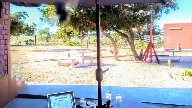 Un espace de restauration en plein air avec une table placée sous un grand parasol, entouré d’une zone sablonneuse avec des arbres et une clôture en arrière-plan.