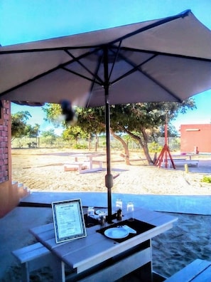 Un área de comedor al aire libre con una mesa bajo un paraguas grande, rodeada por una zona de arena con árboles y una cerca al fondo.