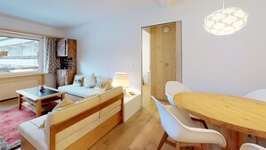 A modern bedroom with a wooden bed, bedside table, and lamp. There is a dining area with a wooden table and chairs.