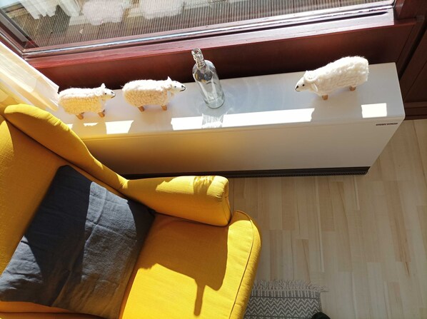 A yellow sofa with a dark cushion, a window with a view of the outside, and a white air conditioning unit with three white sheep figurines on top.