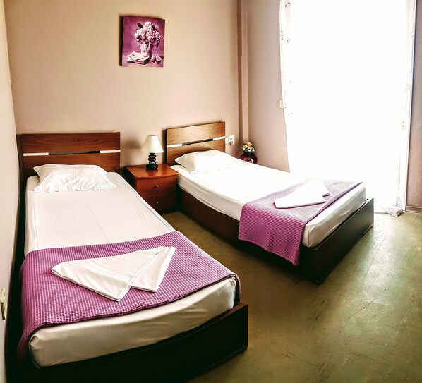 A hotel room with two beds, each with a pink blanket and white linens. There is a nightstand with a lamp and a small vase with flowers. A framed picture hangs on the wall above the beds.
