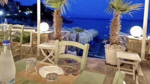 Un espace extérieur aménagé pour les repas, avec des tables et des chaises, une vue sur la mer et des plantes décoratives, notamment des palmiers.