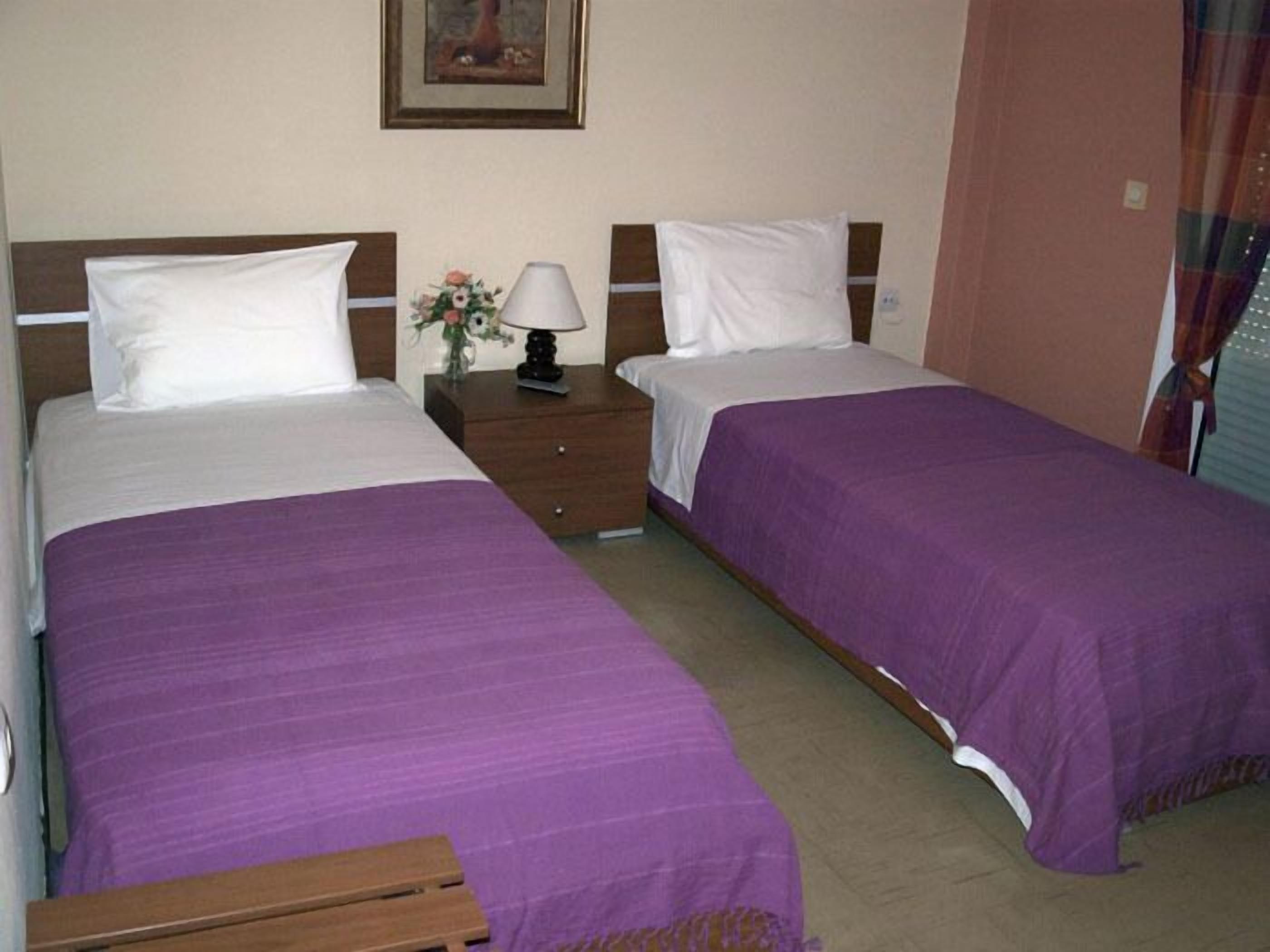 Une chambre d’hôtel avec deux lits simples, chacun recouvert d’une couvre-lit violette et agrémenté de coussins blancs. Une table de chevet avec une lampe et un petit vase de fleurs se trouve entre les deux lits.