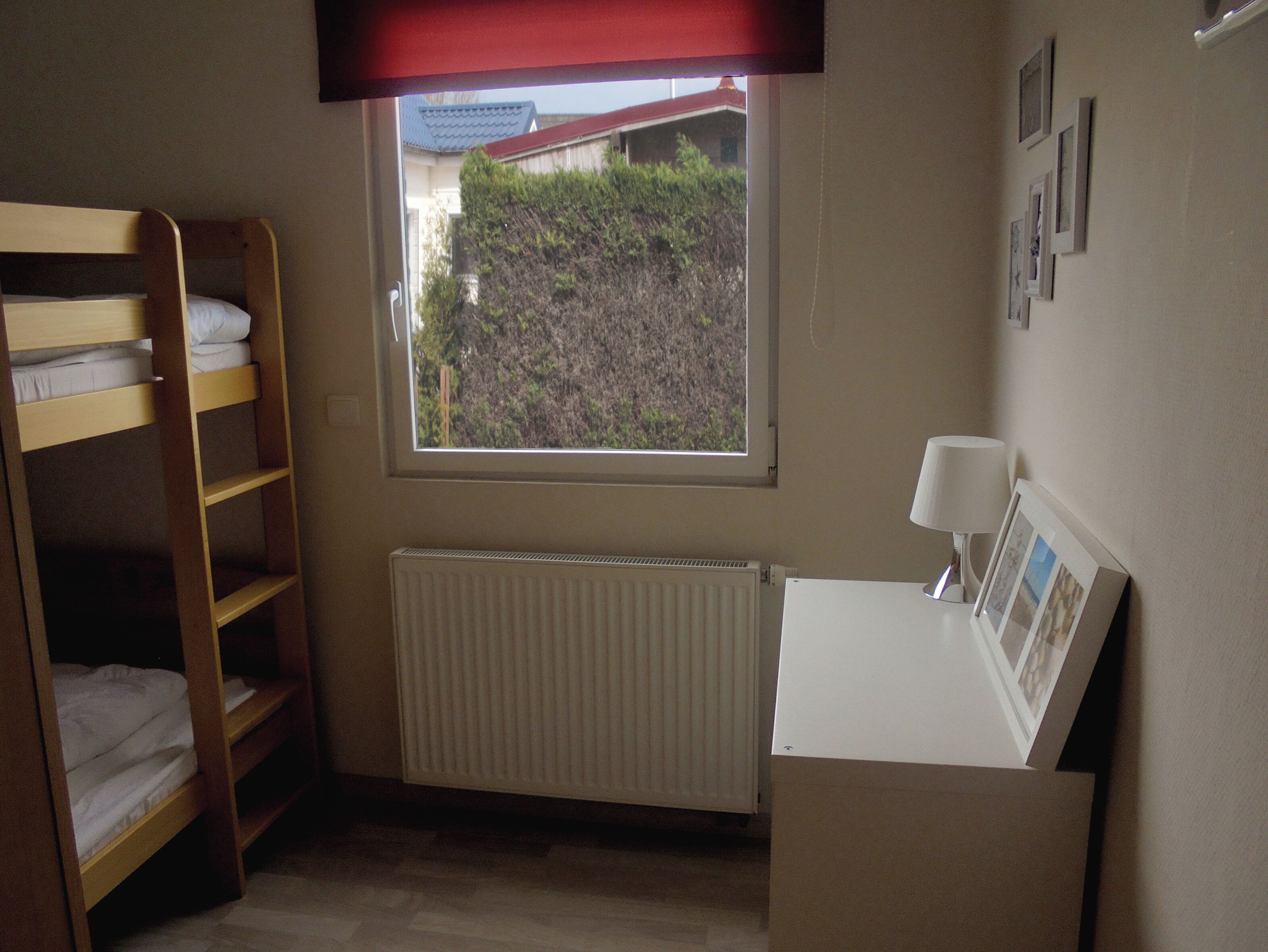 A bedroom with a bunk bed, a radiator, a window with a view of a building and greenery, and a desk with a lamp and framed photos.