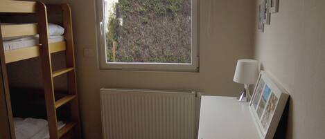 A bedroom with a bunk bed, a radiator, a window with a view of a building and greenery, and a desk with a lamp and framed photos.