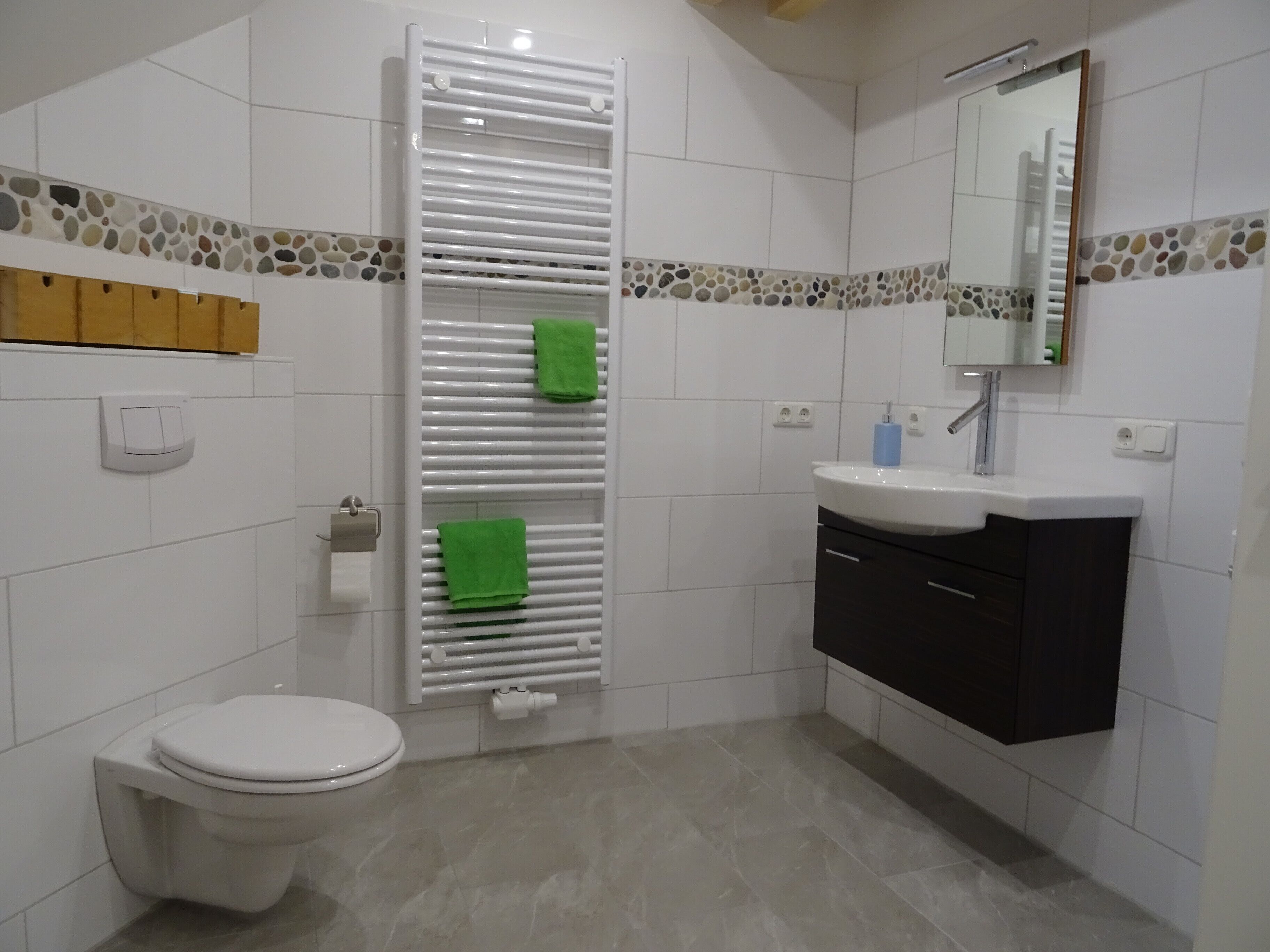 Un bagno moderno con il water bianco, un porta asciugamani con asciugamani verdi, un lavandino con specchio e una cornice a mosaico in pietra.