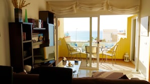 Una sala de estar con sofá, una mesita de centro y una estantería. Hay un balcón con mesa y sillas, y vistas al océano.