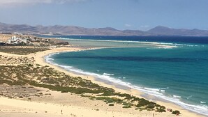 Un paisaje costero con playa de arena, mar azul claro y montañas a lo lejos.