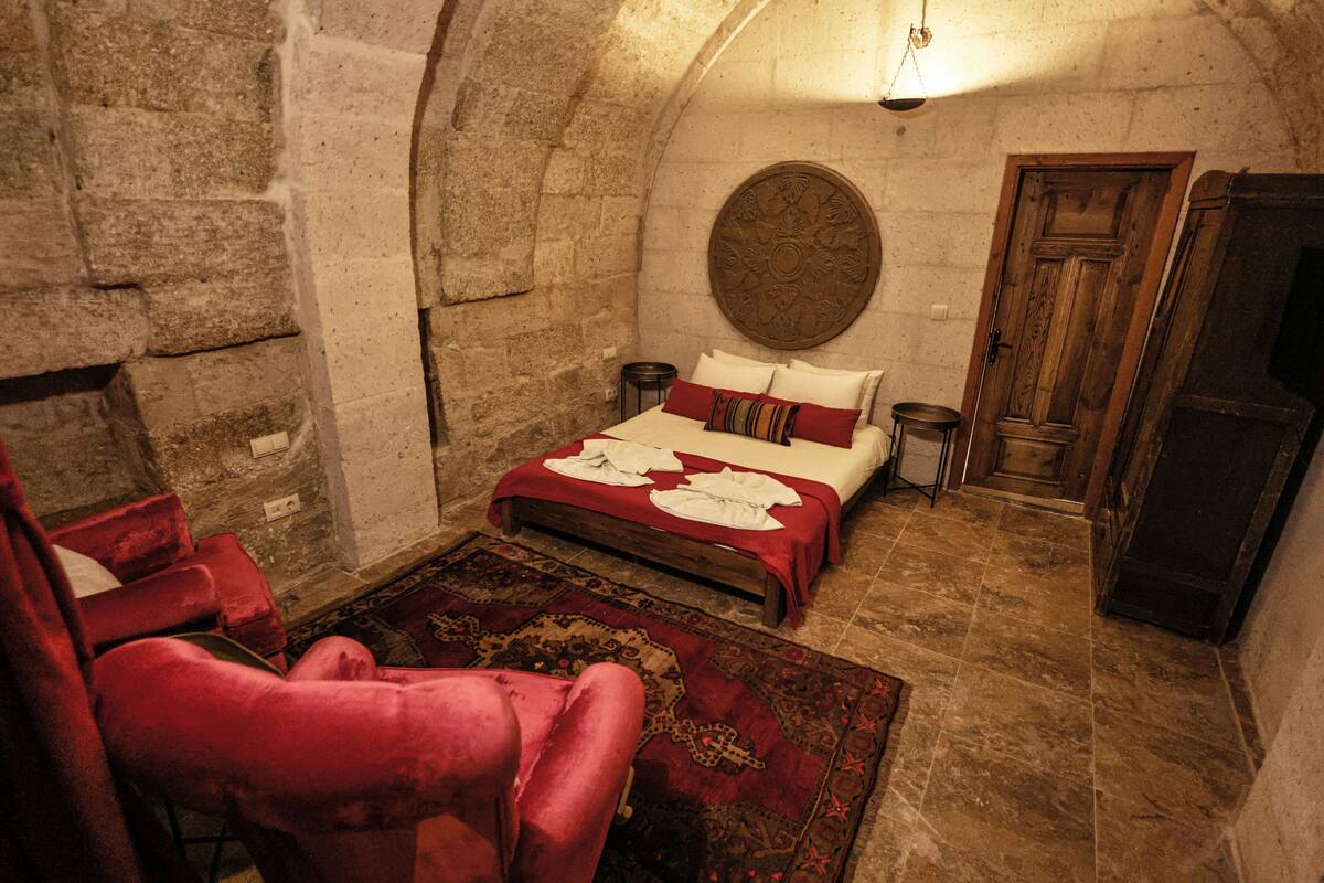 A bedroom with a stone wall, a round decorative plate, a wooden door, a bed with red bedding, and a red armchair.