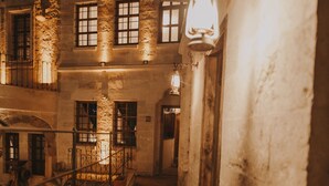 Un estrecho pasillo de paredes de piedra con una escalera que sube a un nivel superior. Se observan varias ventanas con contraventanas, y una lámpara tipo farol se suspende en la pared.